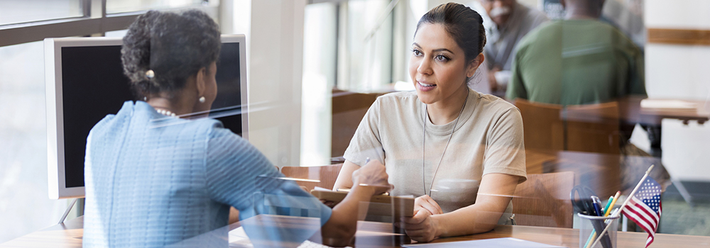Woman talks to another woman seated behind an office desk.